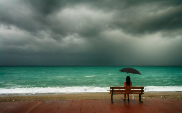 A woman sits alone on a bench by the ocean, holding an umbrella, with a cloudy horizon in the background. The mood is contemplative. This HD desktop wallpaper captures serenity and solitude.