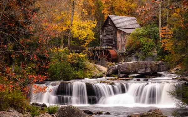 A serene forest scene featuring a flowing river with a cascading waterfall and a rustic watermill nestled among autumn foliage, captured in HD for a desktop wallpaper.