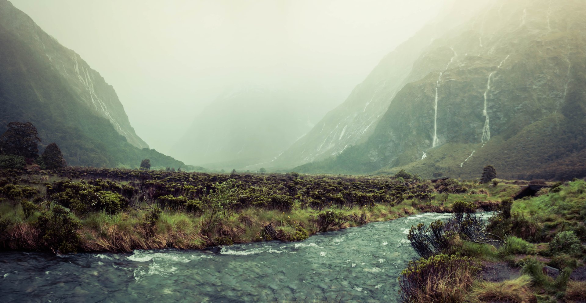 8K Ultra HD PC desktop wallpaper: misty New Zealand valley landscape with a river in the foreground, fog-shrouded hills and cascading waterfalls.