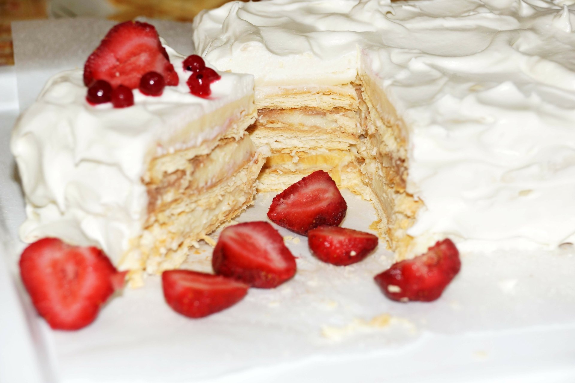 A close-up 4K Ultra HD image of a layered pastry cake with cream and fresh strawberries on a white surface, showcasing its creamy texture and vibrant red fruit.