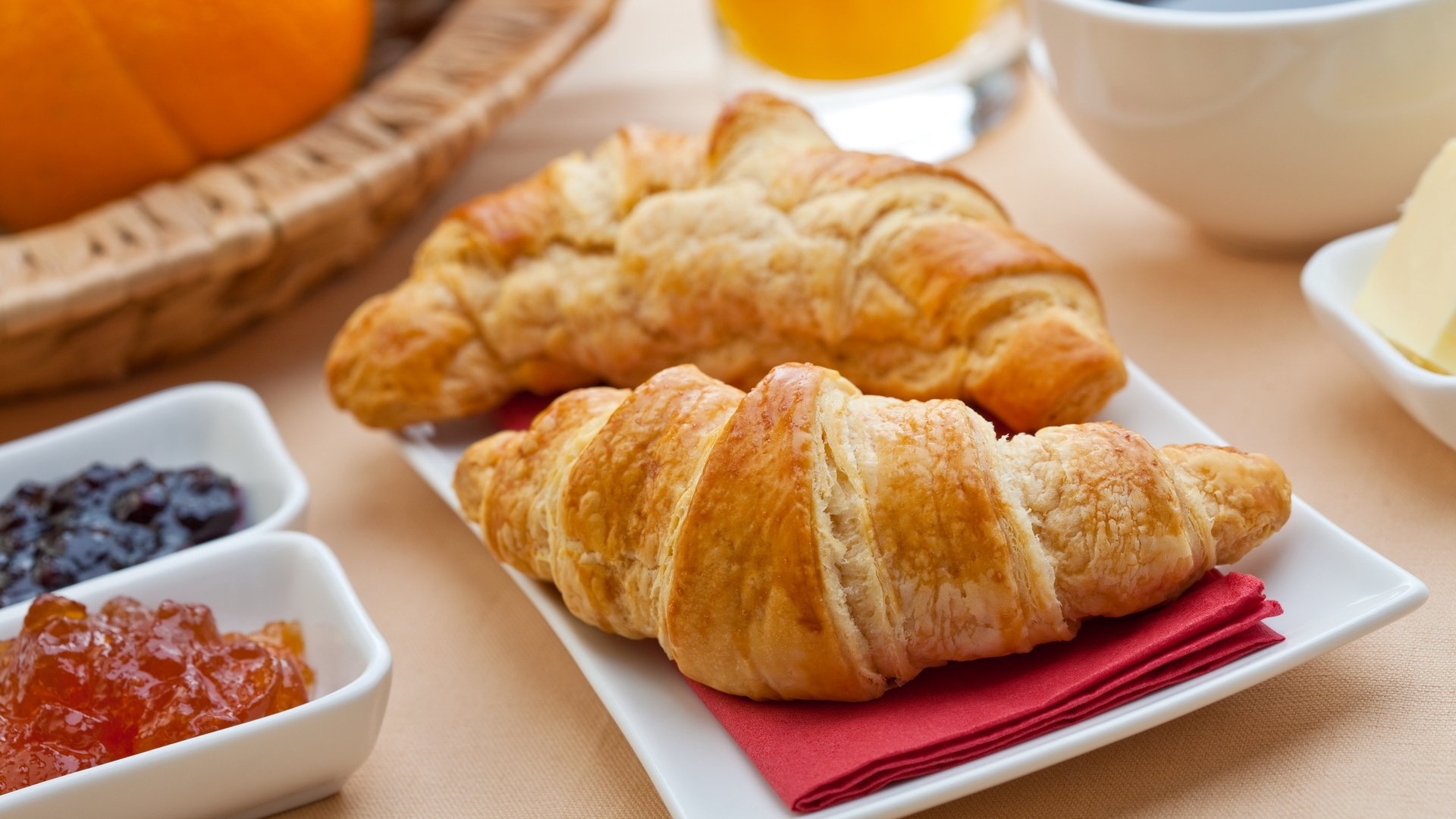 HD desktop wallpaper featuring a close-up of two golden croissants on a white plate, accompanied by breakfast items like jam and orange juice in the background.