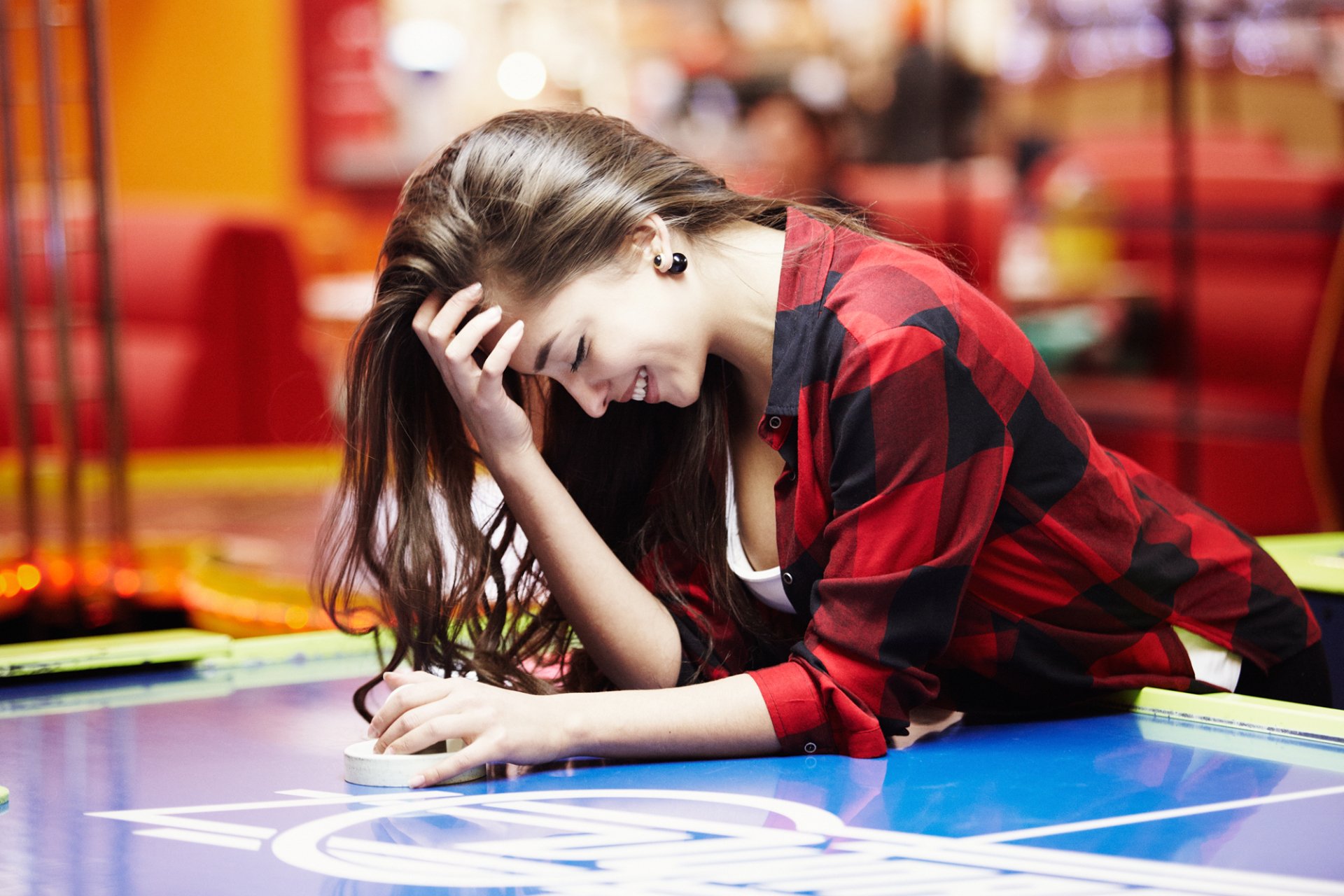 A woman with long hair smiles while leaning on a table, wearing a red plaid shirt. Her relaxed mood captures a carefree moment, set against a vibrant background.