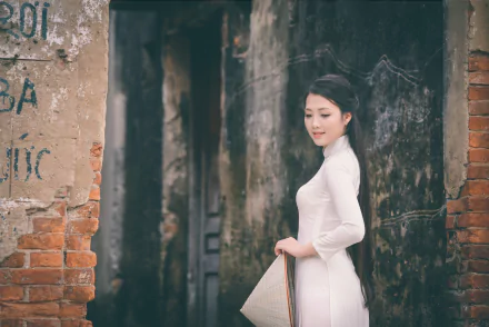 Vietnamese woman wearing a traditional white Ao Dai and an Asian conical hat, posing gracefully against an aged brick and stone wall in a 4K Ultra HD wallpaper.