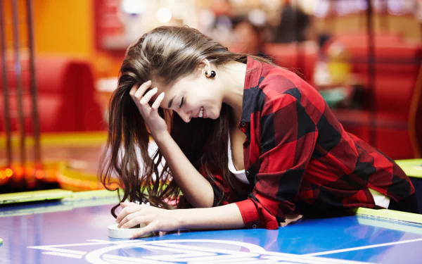 A woman with long hair smiles while leaning on a table, wearing a red plaid shirt. Her relaxed mood captures a carefree moment, set against a vibrant background.