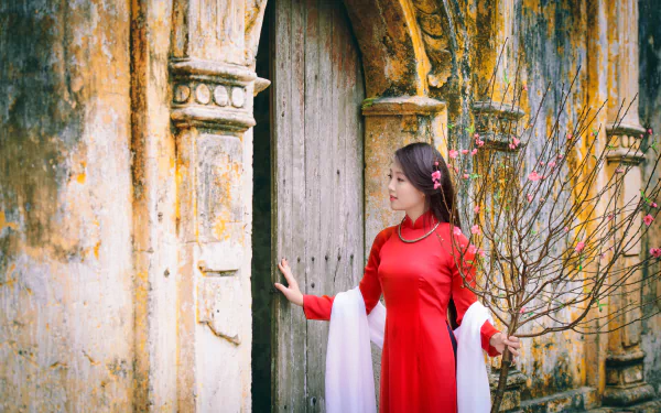 A Vietnamese woman in a red Ao Dai stands beside an aged wooden door and stone archway, set against a textured, weathered wall in this 4K Ultra HD Asian-themed desktop wallpaper.