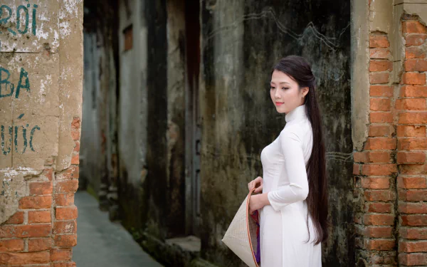 Vietnamese woman in traditional white Ao Dai and Asian conical hat stands in an alley, her long hair flowing, captured in a 4K Ultra HD desktop wallpaper.
