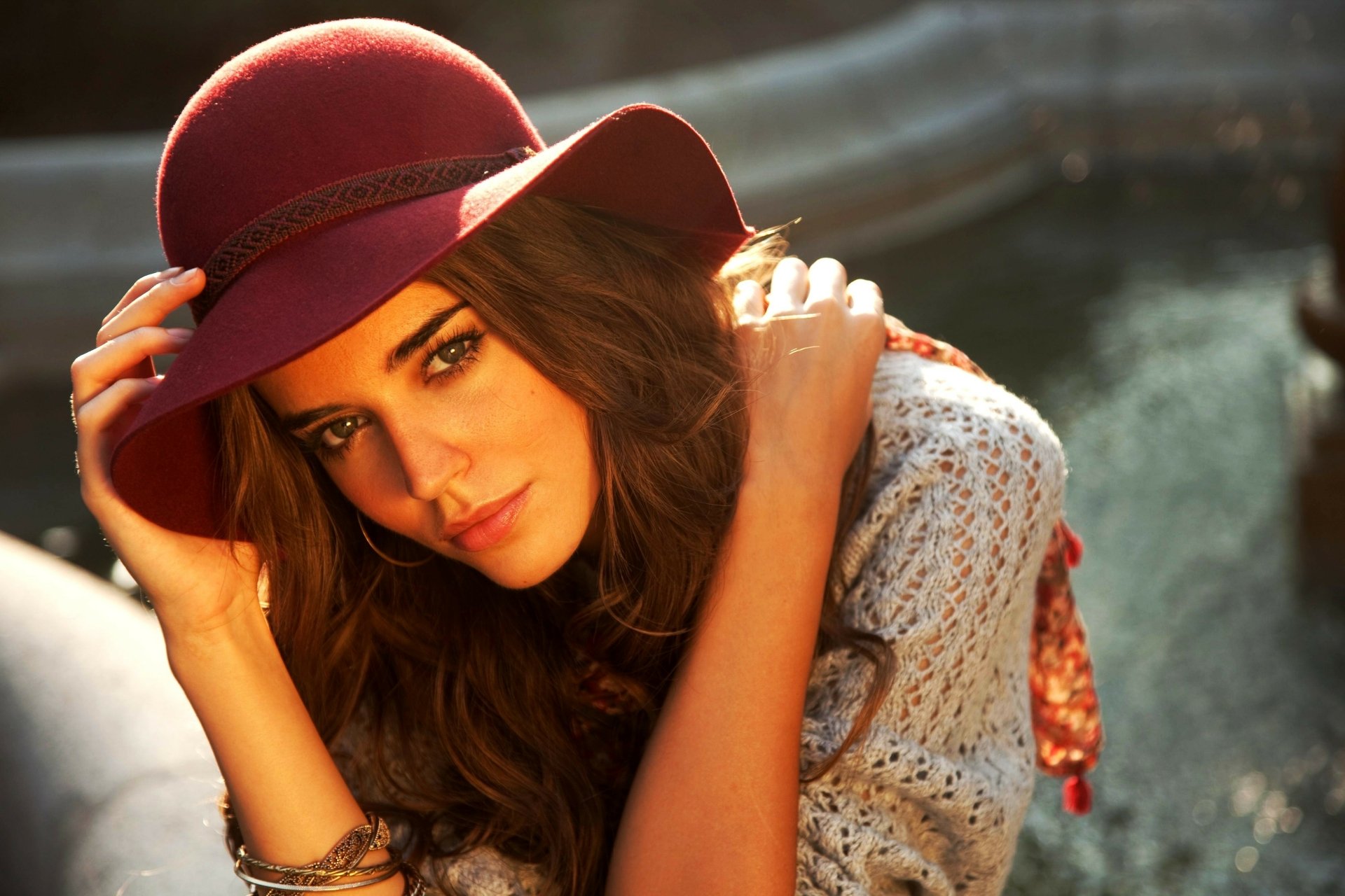 A model with long hair wears a stylish maroon hat, posing gracefully while leaning on her arm, set against a blurred background, creating a captivating 4K Ultra HD wallpaper.