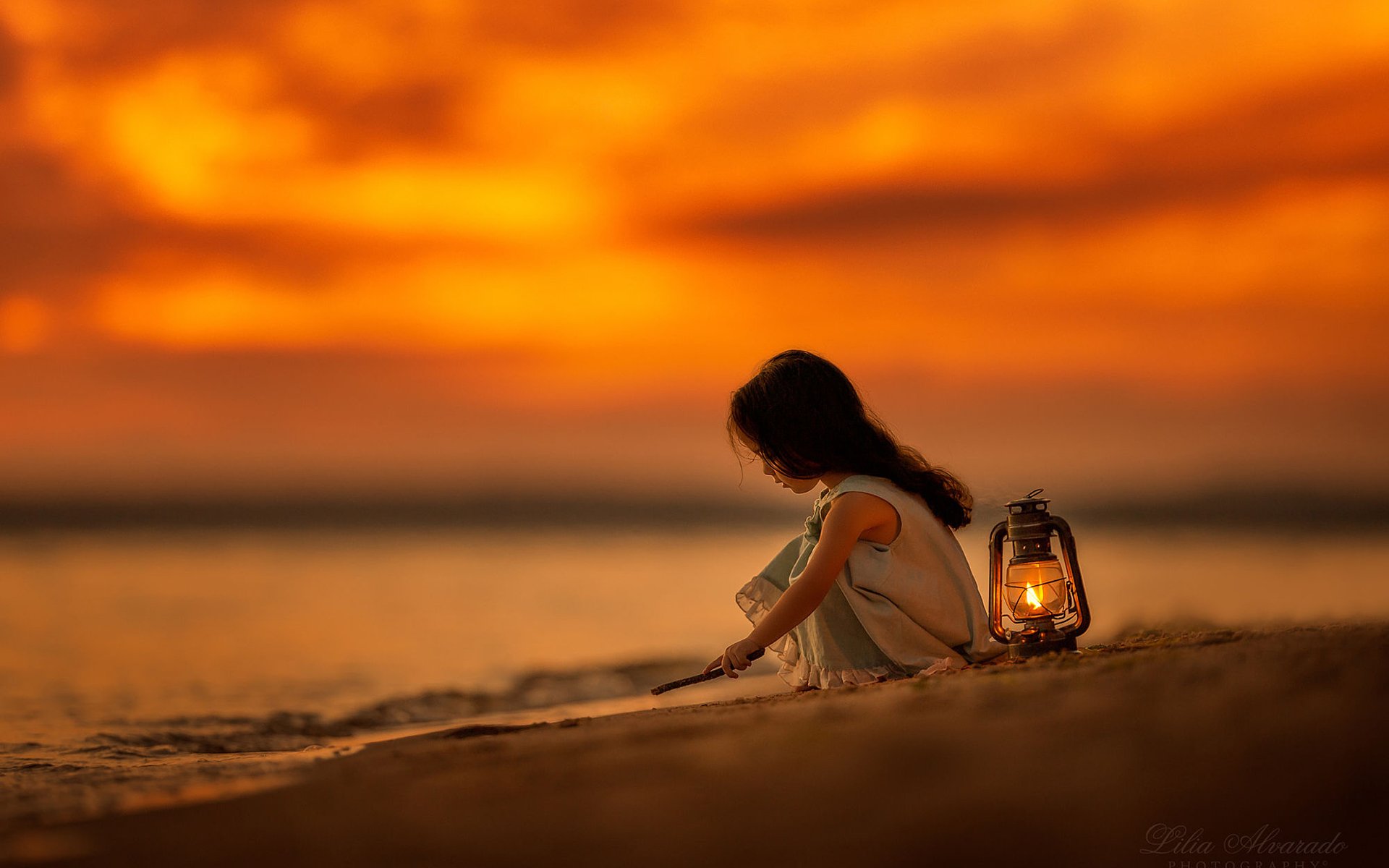 HD PC desktop wallpaper: moody evening beach photograph of a child sitting by the shore with a glowing lantern under a fiery orange sky.