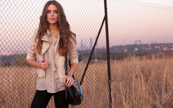 HD desktop wallpaper featuring model Clara Alonso standing in front of a fence in an open field at sunset, wearing a light jacket and carrying a black bag.