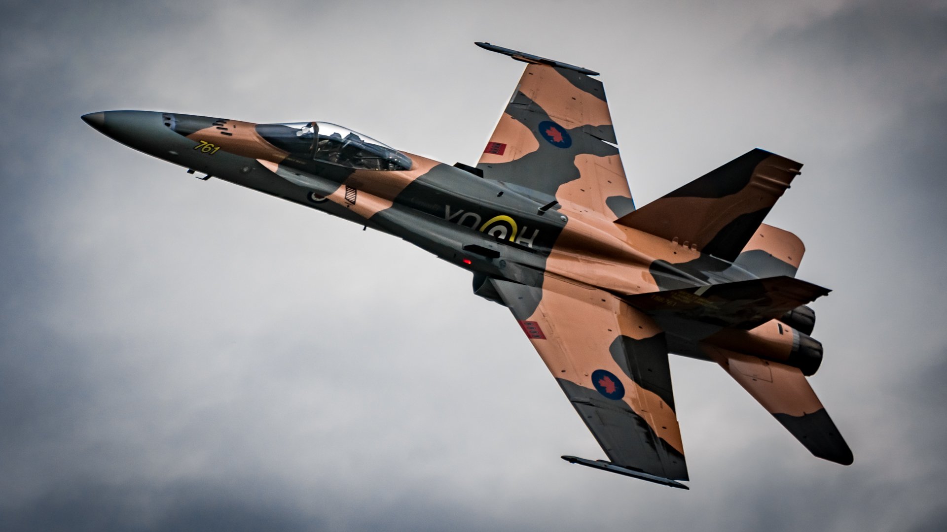 HD desktop wallpaper of a McDonnell Douglas CF-18 Hornet jet fighter warplane in military camouflage flying against a cloudy sky.