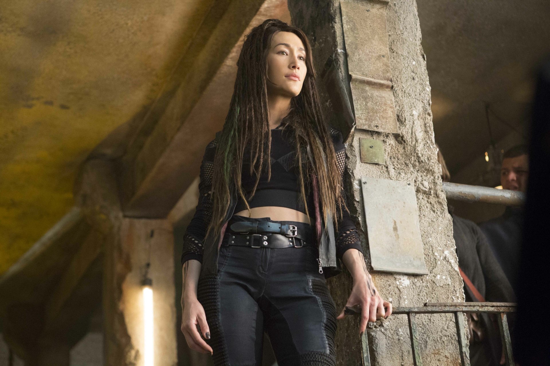 Maggie Q as Tori from The Divergent Series: Allegiant stands confidently in a dimly lit industrial setting, captured in a 4K Ultra HD movie wallpaper.