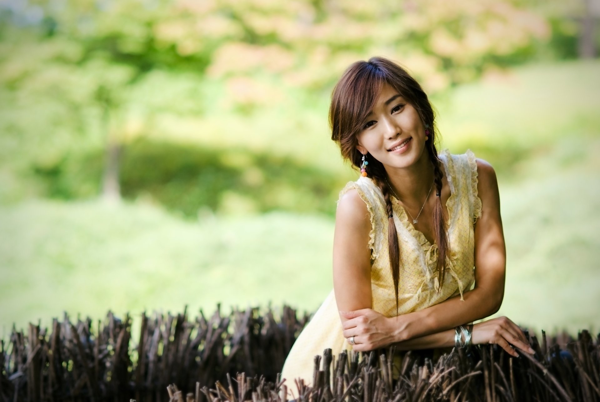 A smiling Asian woman with brown eyes and a braid poses gracefully outdoors, surrounded by a soft bokeh background, making this a captivating 4K Ultra HD wallpaper.