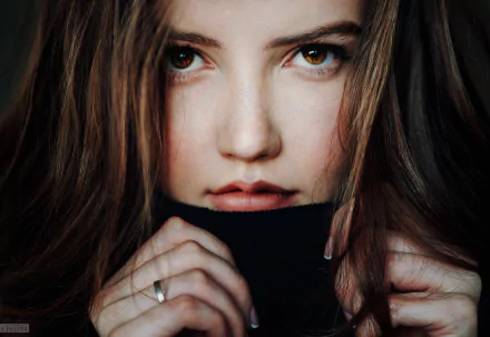 A close-up of a model with brown eyes and brunette hair, gently pulling a dark sweater collar around her face, conveying a captivating and serene expression. HD desktop wallpaper.