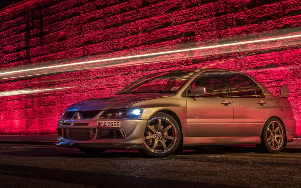 A Mitsubishi Lancer Evolution is showcased in a striking HD desktop wallpaper, illuminated by vibrant red lighting against a textured wall, highlighting its sleek design and sporty features.