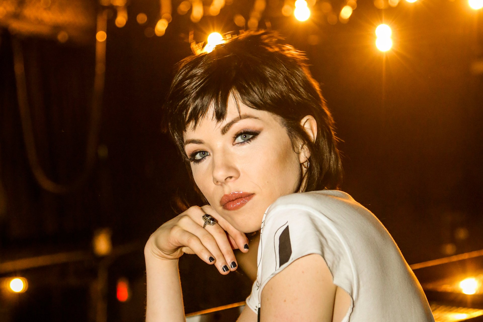 Blue-eyed brunette woman gazing at the camera with warm stage lights behind her — HD music-themed desktop wallpaper.