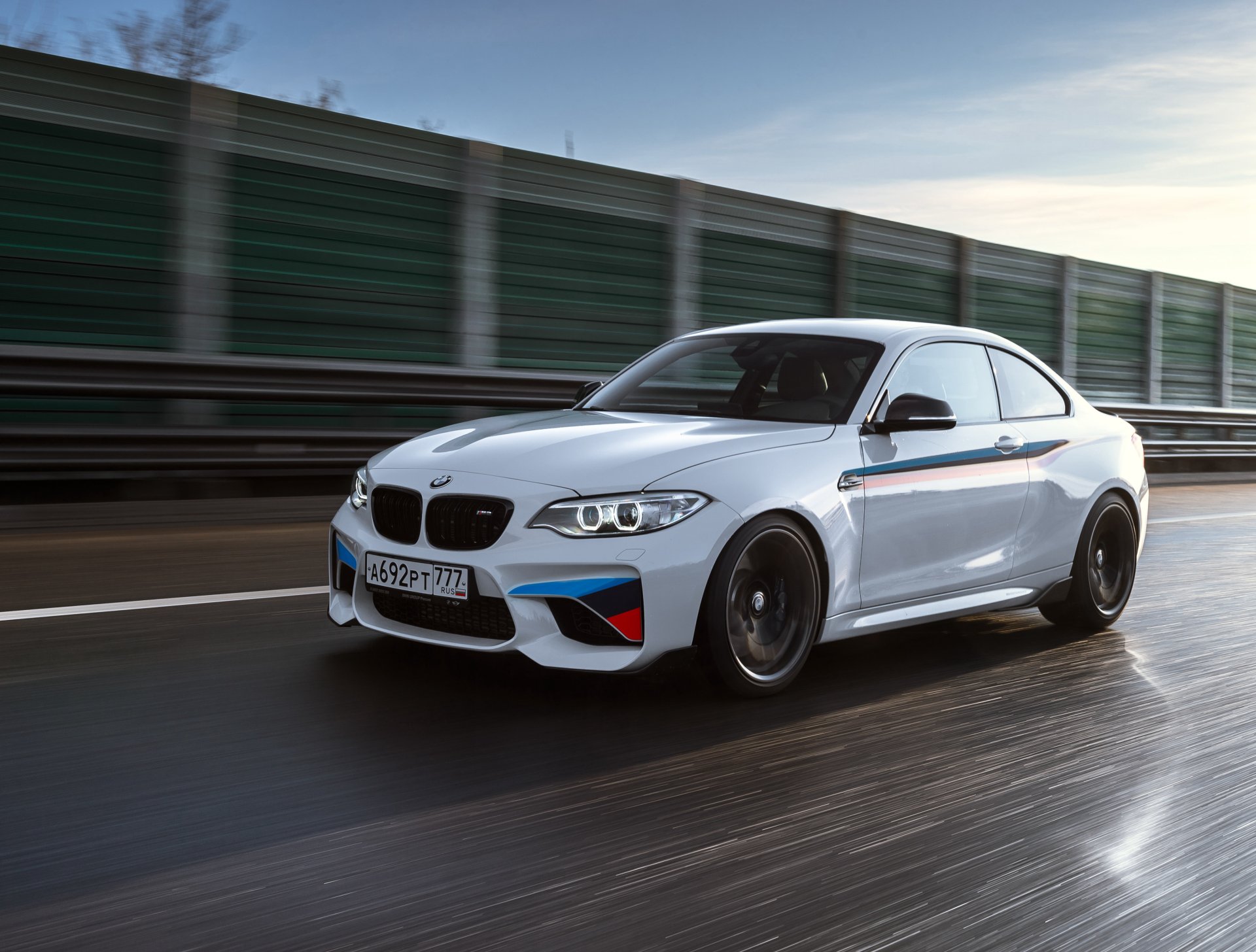 White BMW M2 sports car driving on a wet road, captured in sharp 4K Ultra HD quality, showcasing the vehicle's sleek design and dynamic motion.
