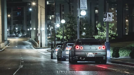 Rear view of two Nissan Skyline GT-R R34 vehicles parked on a wet city street at night, neon streetlights reflecting on the pavement — HD PC desktop wallpaper and background.