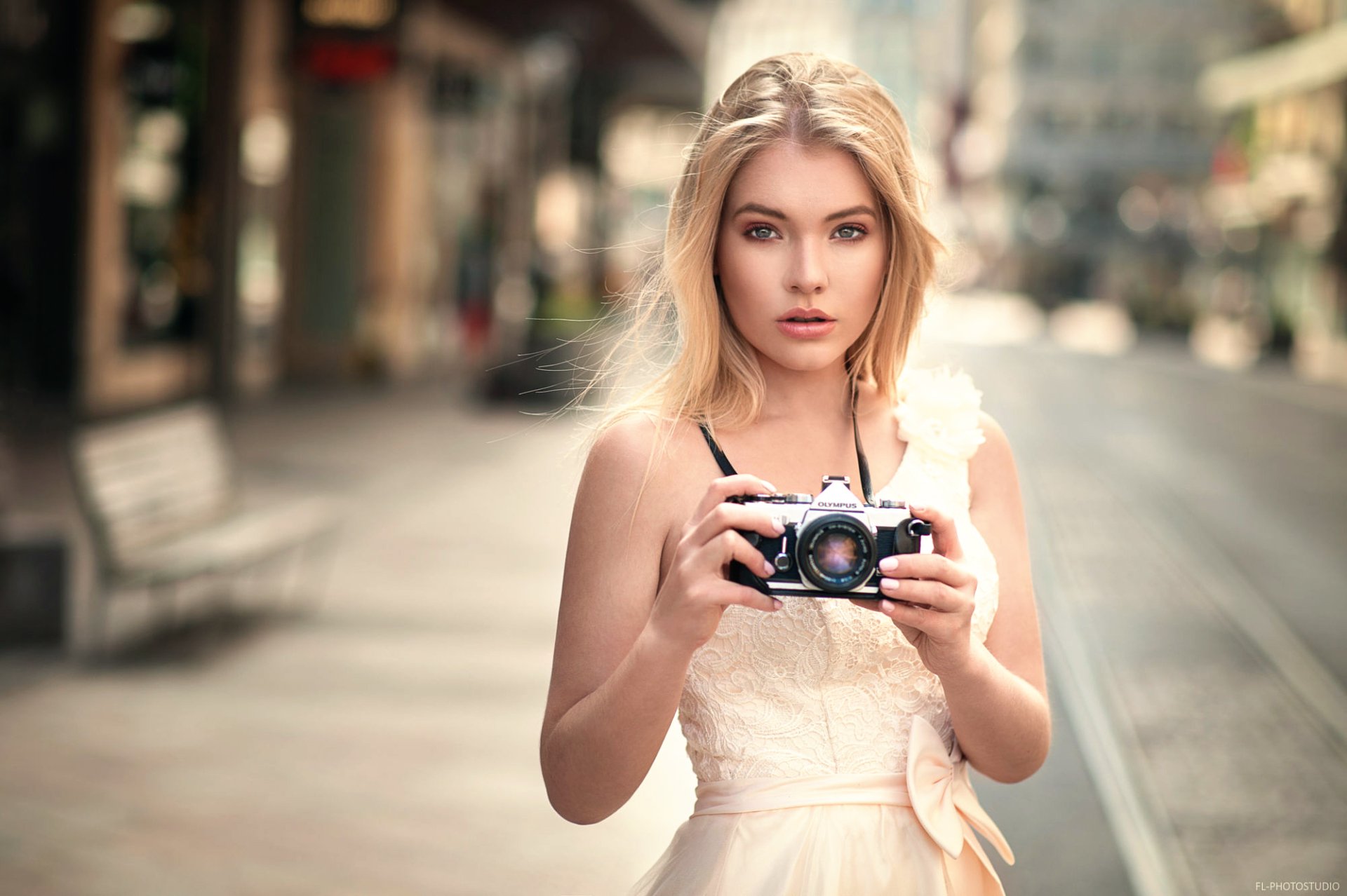 A blonde woman with long hair holds an Olympus camera while standing on a blurred street. She wears a light dress and looks at the camera, creating a stunning HD desktop wallpaper and background.