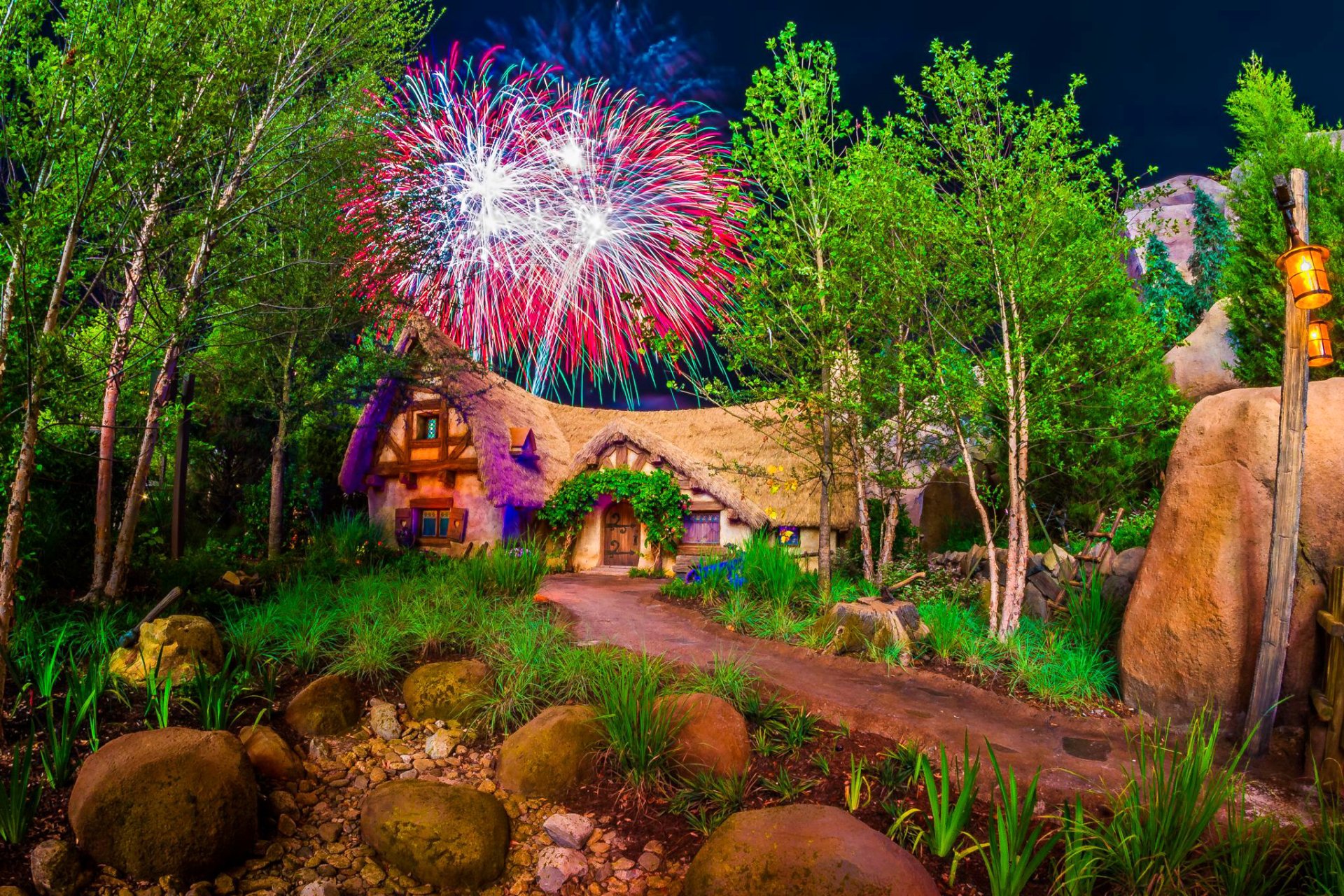 HD desktop wallpaper showing a charming house at Walt Disney World's Disneyland with vibrant fireworks lighting up the night sky above the lush, green surroundings.