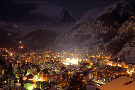 A snowy winter night in a Swiss Alpine village nestled in a mountain valley, illuminated by warm lights against dark peaks under a night sky.