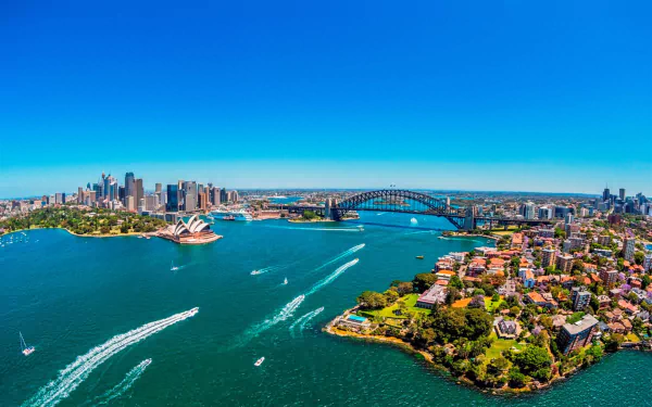 Aerial view of Sydney Harbour showcasing the Sydney Opera House, Sydney Harbour Bridge, city skyline, and surrounding ocean under a clear blue sky.