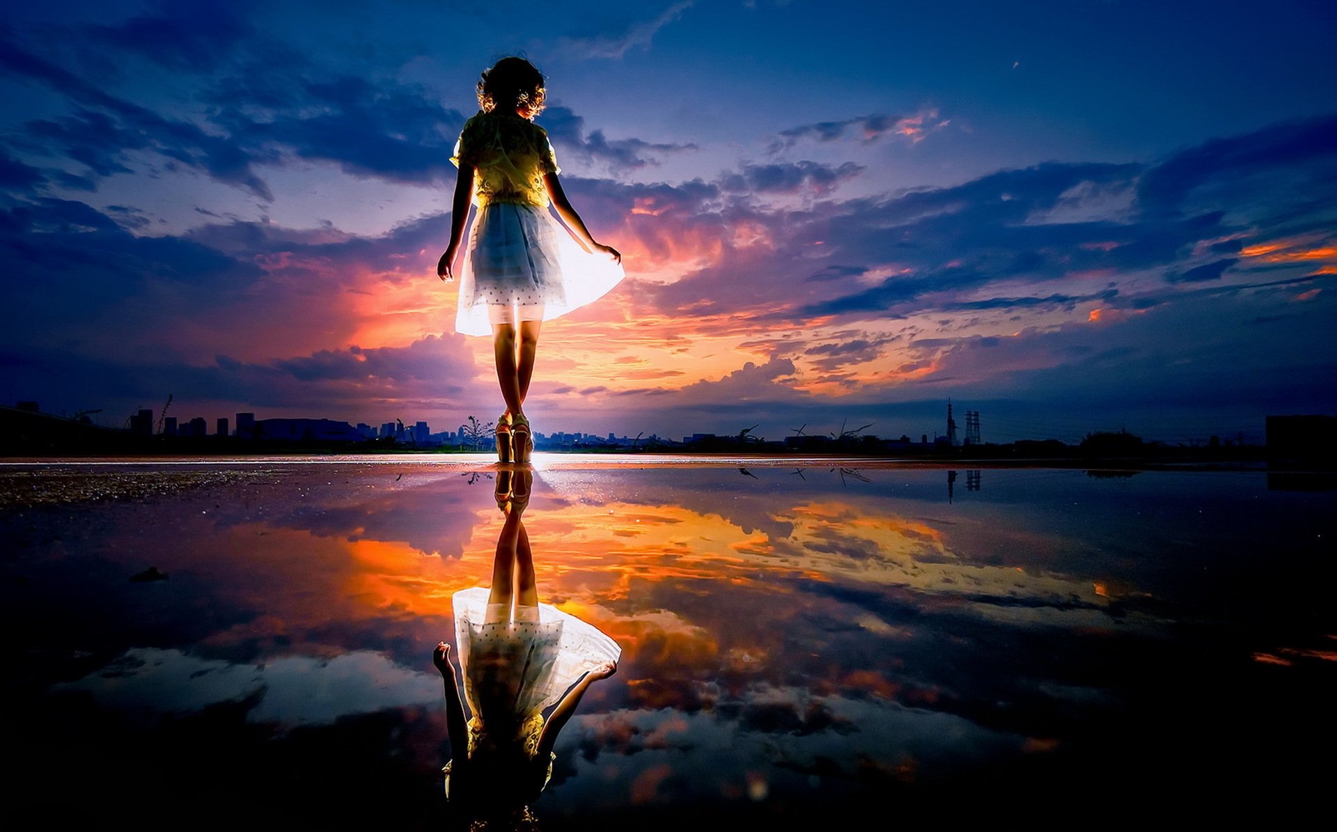 A silhouetted woman stands gracefully in a puddle, reflecting a vibrant sunset sky filled with dramatic clouds, creating an artistic and serene atmosphere.