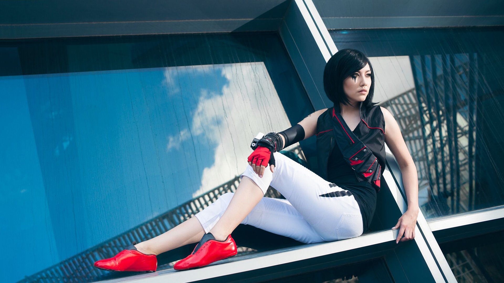 A woman in brunette cosplay as Faith Connors from Mirror's Edge poses confidently against a reflective backdrop, showcasing her dynamic outfit and vibrant red accessories.