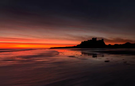 Silhouette of Bamburgh Castle on the beach at sunset with an orange horizon, captured in England as a stunning HD desktop wallpaper.