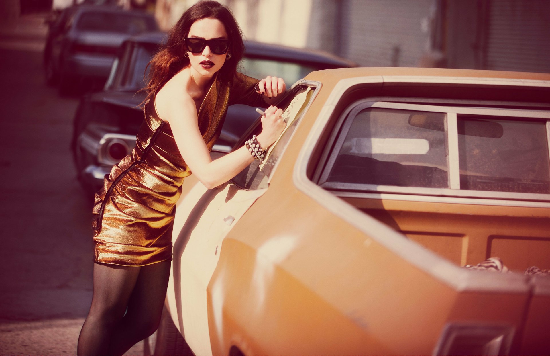 Brunette woman in a gold mini dress, red lipstick and sunglasses leaning on an orange vintage car — 2K Quad HD desktop wallpaper.