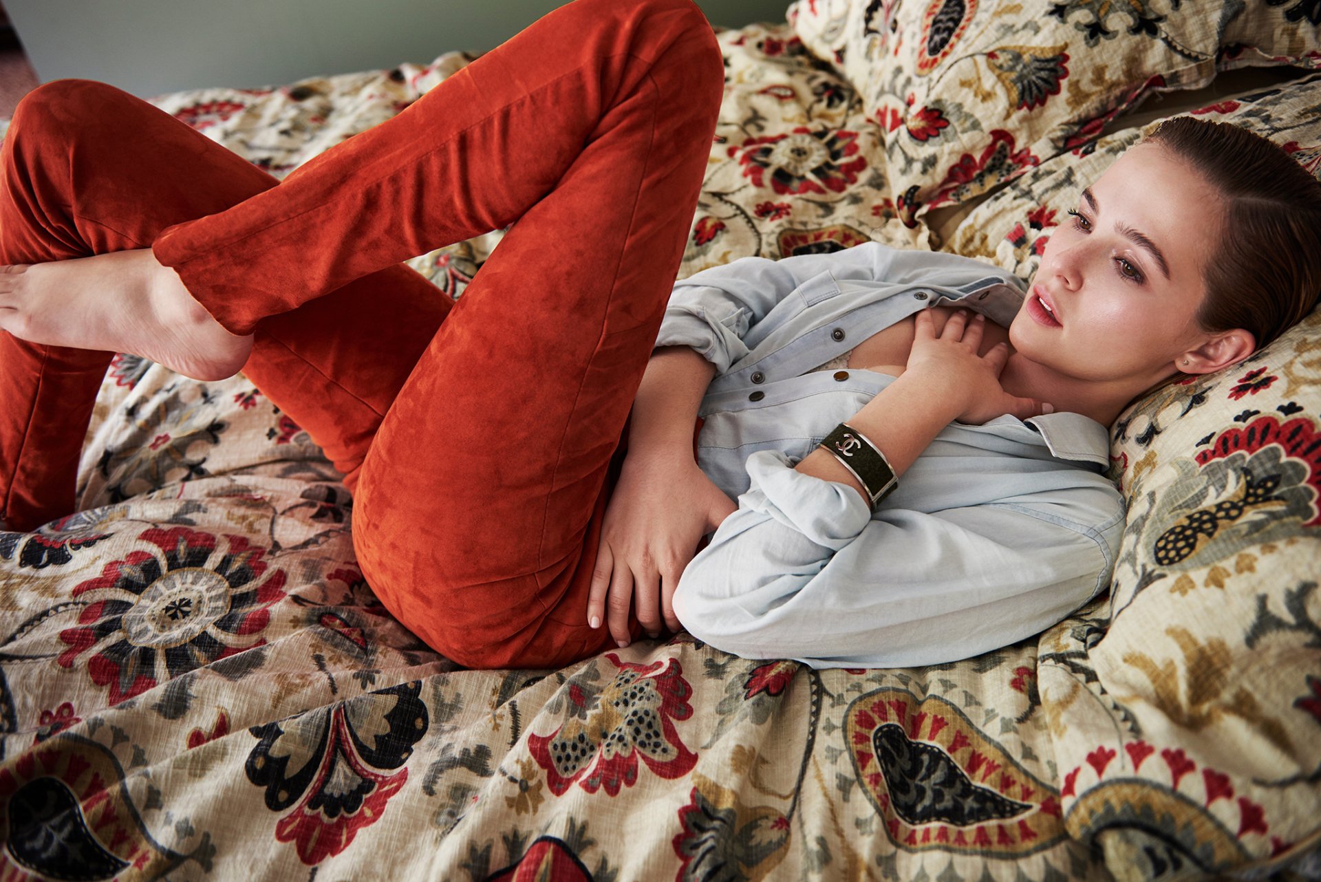HD desktop wallpaper featuring American actress Zoey Deutch with brown eyes and brunette hair, reclining on a patterned sofa wearing rust-colored pants and a light shirt.