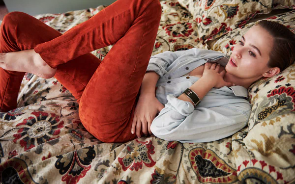 HD desktop wallpaper featuring American actress Zoey Deutch with brown eyes and brunette hair, reclining on a patterned sofa wearing rust-colored pants and a light shirt.