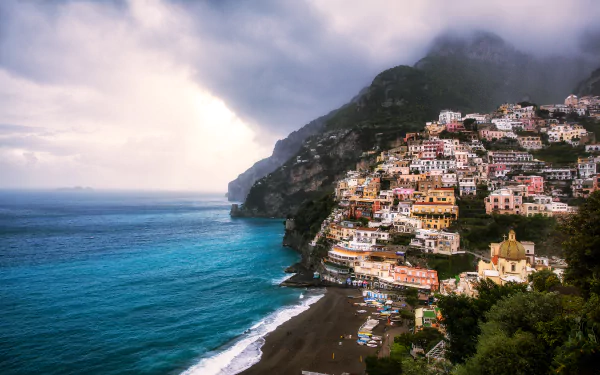 HD desktop wallpaper of Positano, Italy, showcasing the colorful town cascading down the coastline beside a calm beach under a cloudy sky.