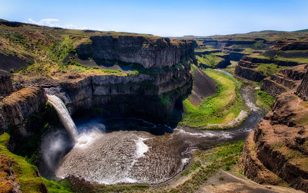 A stunning HD landscape of Palouse Falls cascading into a river winding through a rugged canyon under a clear blue sky.