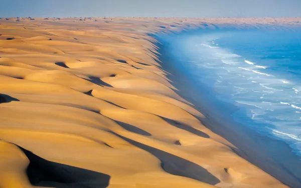 HD desktop wallpaper of Namibia's desert dunes meeting the Atlantic Ocean — golden sand slopes beside blue sea under a clear sky, an evocative nature coastline.