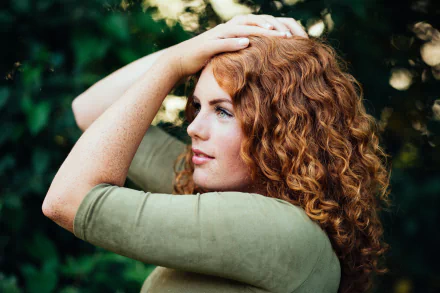 A woman with vibrant red curls and green eyes poses gracefully, her freckles lightly accentuated. The background features a soft bokeh effect, enhancing the mood of the image.