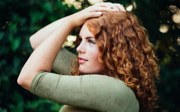 A woman with vibrant red curls and green eyes poses gracefully, her freckles lightly accentuated. The background features a soft bokeh effect, enhancing the mood of the image.