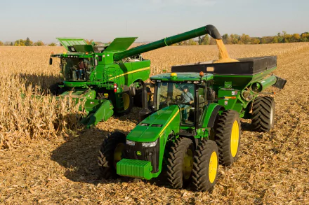 A vibrant HD desktop wallpaper featuring John Deere vehicles working in a cornfield, showcasing a combine harvester and tractor in action amidst golden crops.