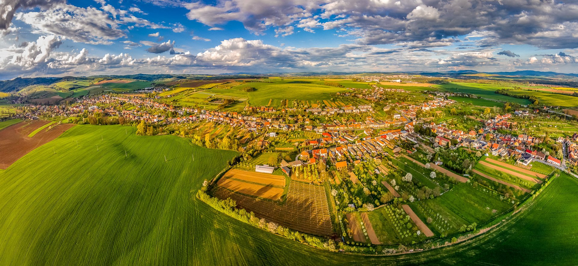 A panoramic aerial view of a Czech Republic landscape featuring expansive green fields, a small town under a vast sky with scattered clouds, captured in HD photography.