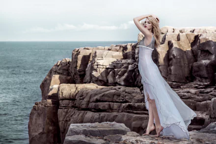 A blonde woman model stands on rocky cliffs wearing a flowing white dress against a serene ocean backdrop, captured in an 8K Ultra HD PC desktop wallpaper.