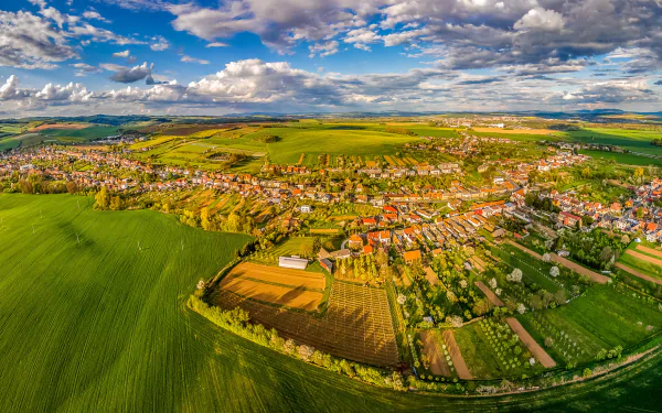 A panoramic aerial view of a Czech Republic landscape featuring expansive green fields, a small town under a vast sky with scattered clouds, captured in HD photography.