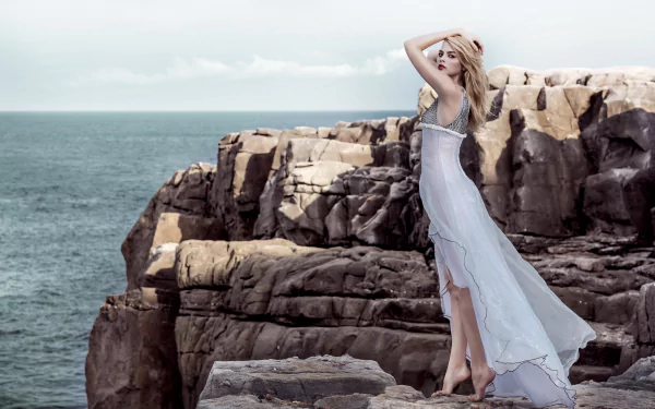 A blonde woman model stands on rocky cliffs wearing a flowing white dress against a serene ocean backdrop, captured in an 8K Ultra HD PC desktop wallpaper.