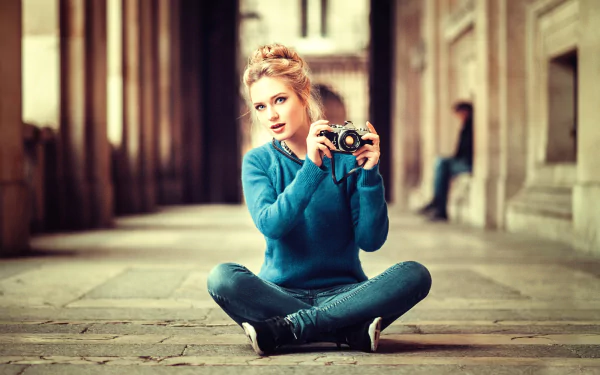 A blonde model with blue eyes sits on the ground, holding a camera, dressed in a blue sweater and jeans, surrounded by an architectural backdrop.