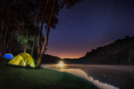 A tranquil camping scene features illuminated tents beside a serene lake, framed by a lush forest under a starry night sky, creating a captivating 4K Ultra HD desktop wallpaper.