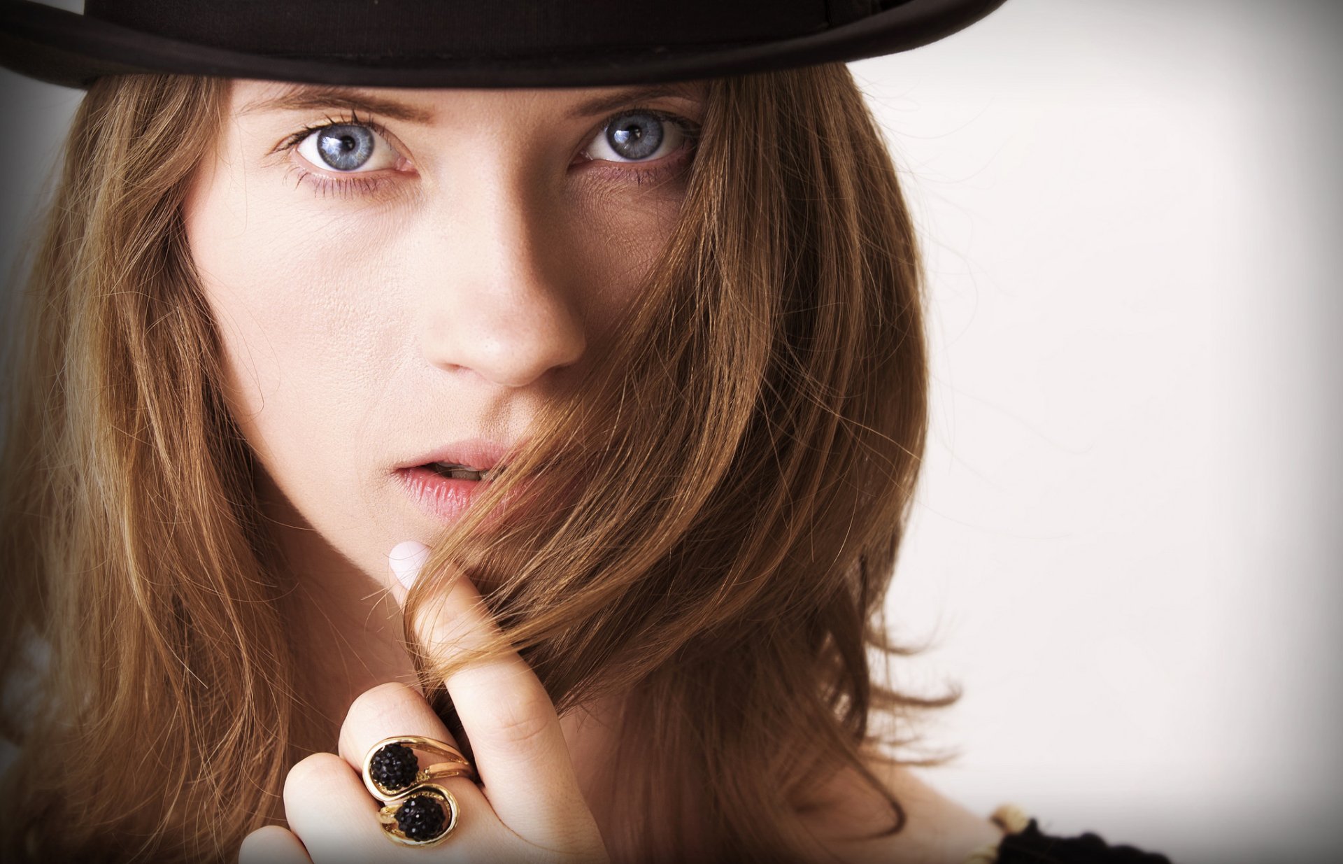 Close-up portrait of a brunette woman with blue eyes wearing a black hat, holding her hair near her face, showcasing a ring on her finger in this HD desktop wallpaper.