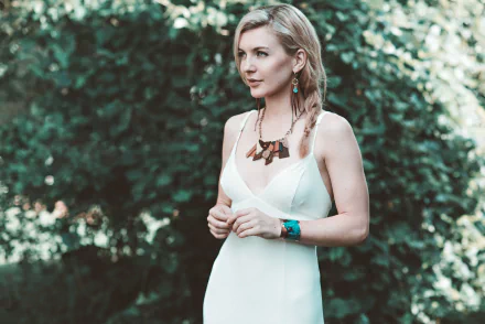 Blonde woman with green eyes wearing a white dress, earrings, and necklace, her hair in a braid, standing in front of a blurred green bokeh background.