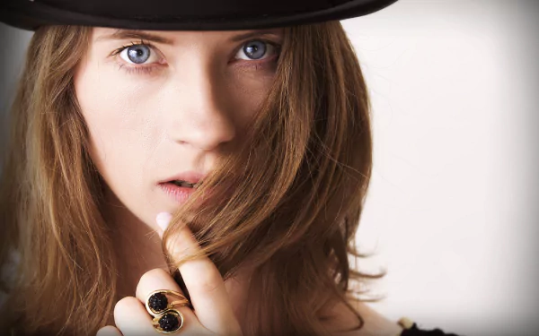 Close-up portrait of a brunette woman with blue eyes wearing a black hat, holding her hair near her face, showcasing a ring on her finger in this HD desktop wallpaper.