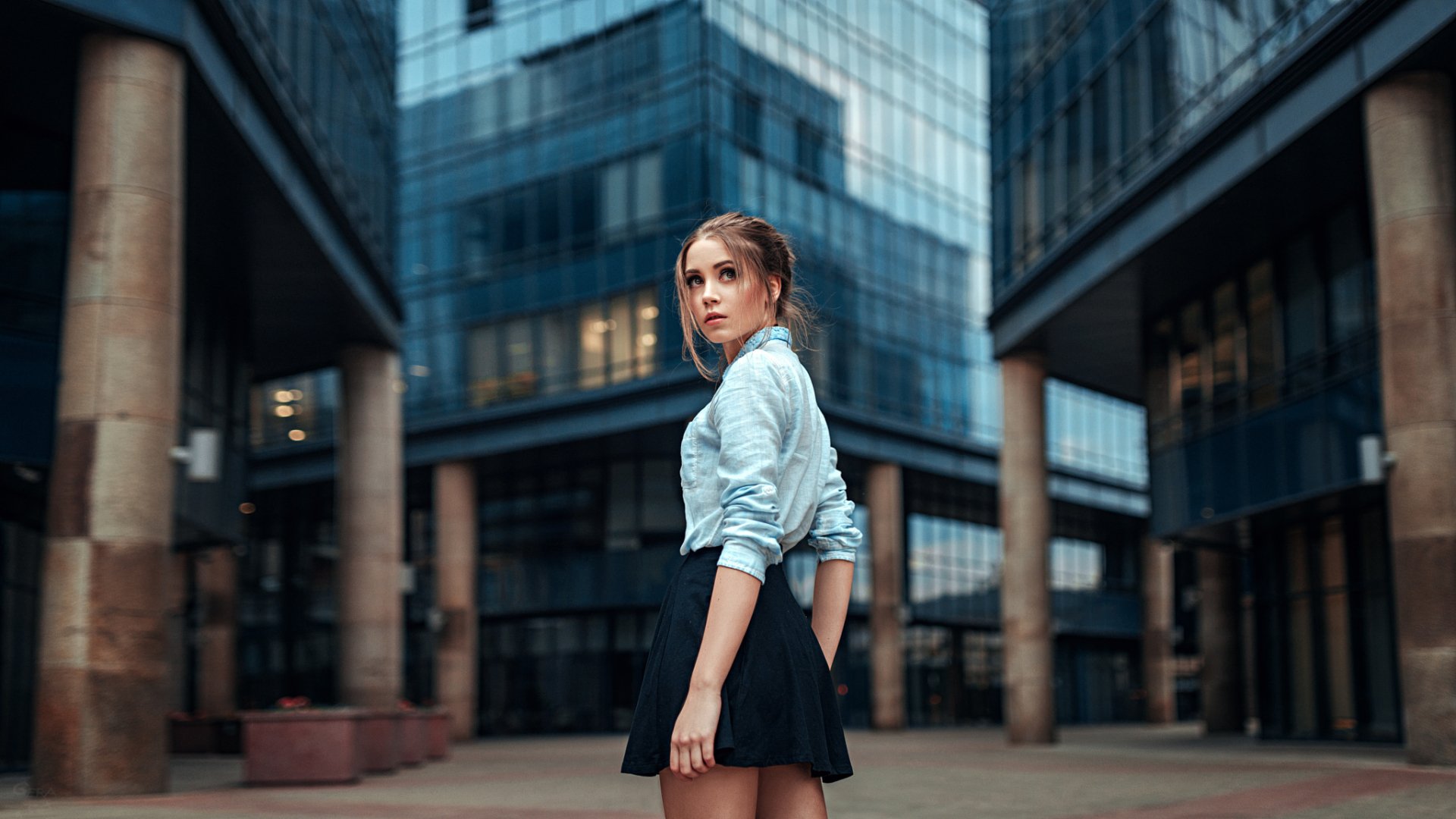 A HD desktop wallpaper featuring a brunette woman in a skirt named Kseniya Kokoreva, standing in an urban setting with modern glass buildings in the background.