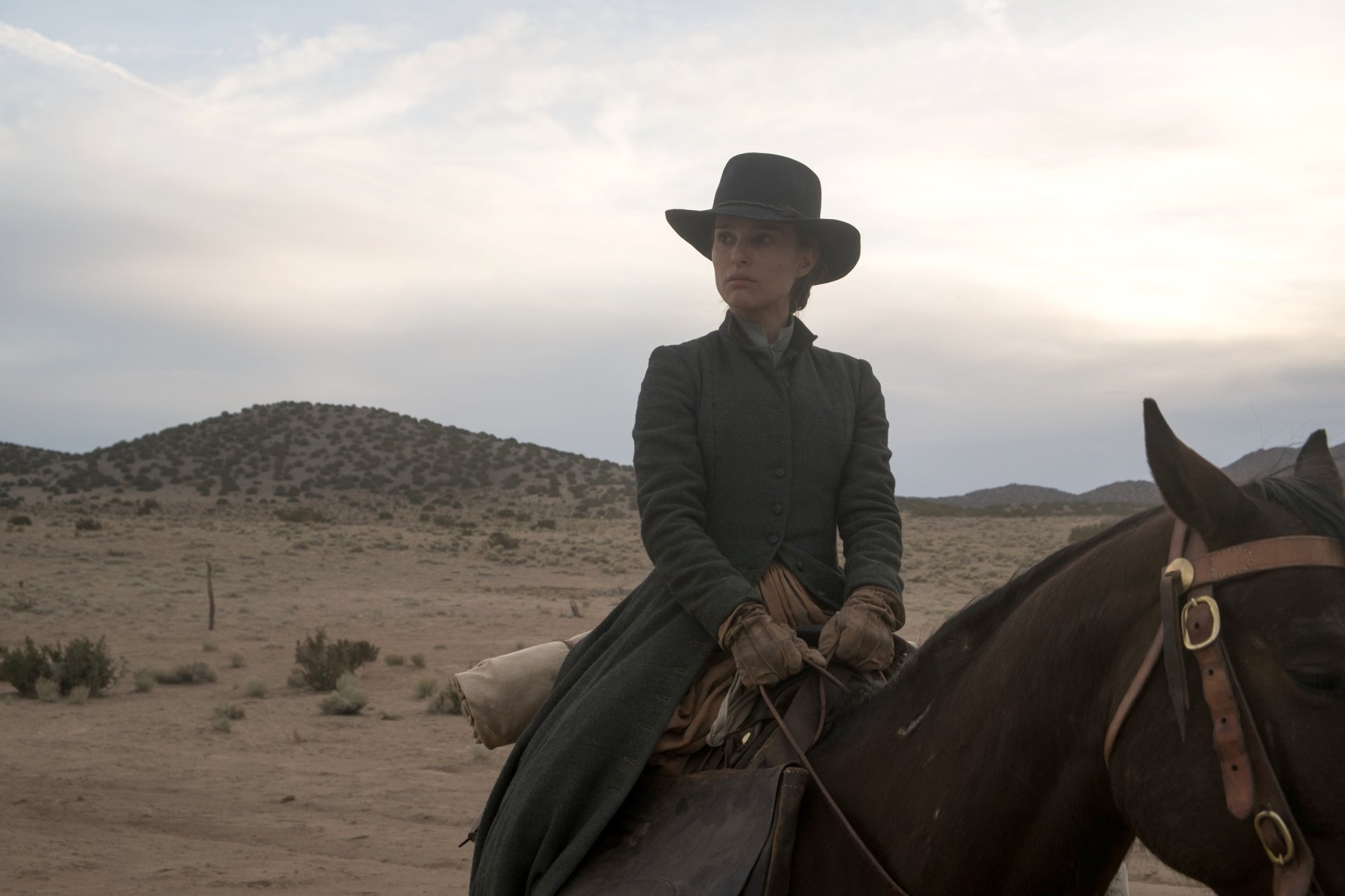 4K Ultra HD desktop wallpaper: woman in a dark coat and wide-brim hat on horseback crossing a desert, cinematic scene from the film Jane Got a Gun.