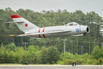 HD PC desktop wallpaper: military Mikoyan-Gourevitch MiG-17 jet, white with red markings and tail star, flying low above runway with dense green forest backdrop.