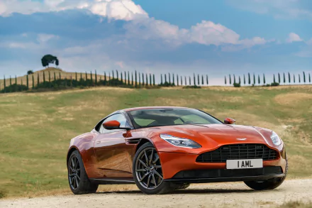 Orange Aston Martin DB11 grand tourer parked on a dirt road with rolling hills and a partly cloudy sky in the background, presented as a 4K Ultra HD desktop wallpaper.
